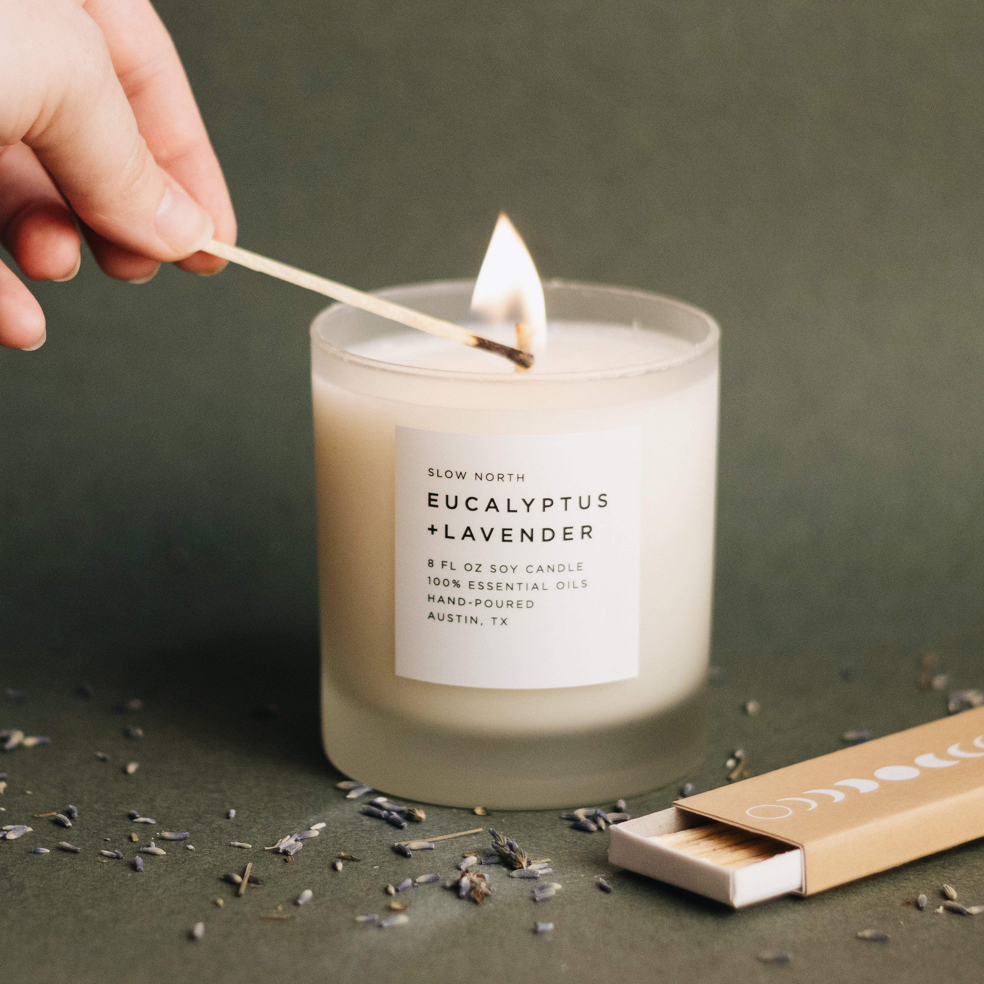 hand lighting white soy wax candle with Eucalyptus + Lavender on the label