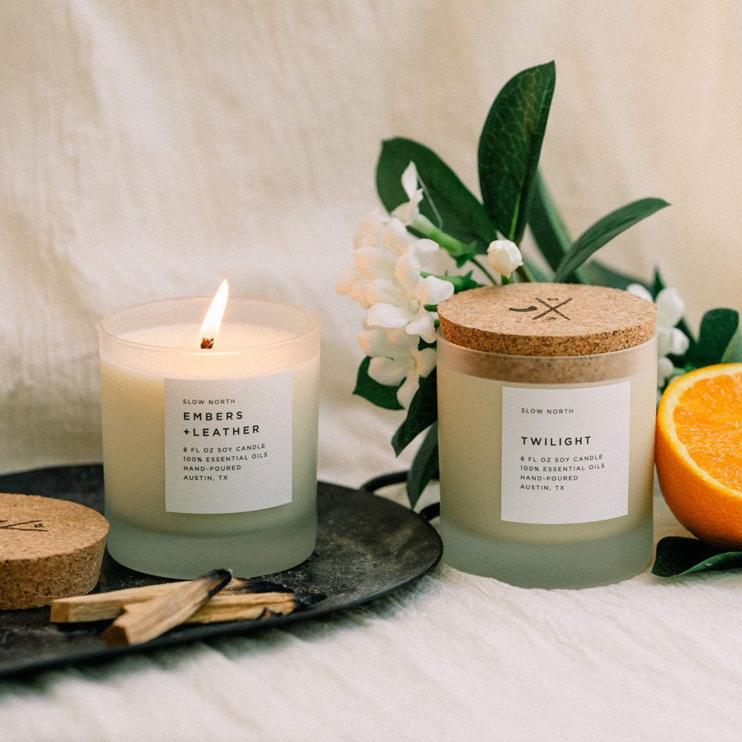 Embers + Leather and Twilight by Slow North on a white background, surrounded by palo santo sticks, a sliced orange, and florals.